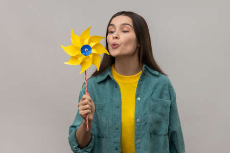 Hand mill. Portrait of happy beautiful young adult woman blowing at paper windmill, pinwheel toy on...
