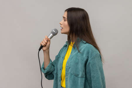 Side view portrait of young adult woman journalist holding microphone, discussing problems, reporting, wearing casual style jacket. Indoor studio shot isolated on gray background.の写真素材