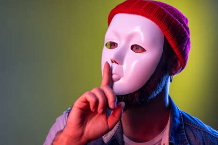 Side view of unknown anonymous man in white mask on face, hiding personality, keeping finger near...