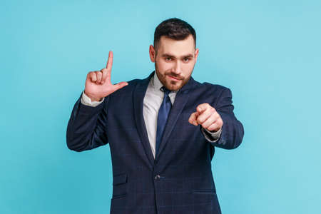 You lost. Portrait of dissatisfied businessman with beard in suit showing loser or lame gesture and pointing to camera, accusing for failure. Indoor studio shot isolated on blue background.の写真素材
