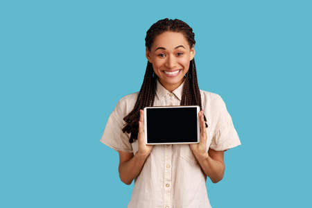 Smiling satisfied woman holds modern tablet with empty screen for your advertisement, poses with electronic gadget in hands, wearing white shirt. Indoor studio shot isolated on blue background.の写真素材