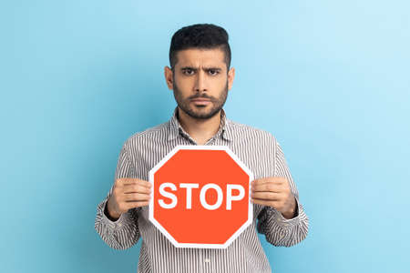 Portrait of serious bearded businessman holding Stop road traffic sign as symbol of prohibition, restrictions, looking at camera, wearing striped shirt. Indoor studio shot isolated on blue background.の写真素材
