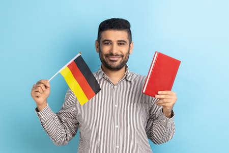 Portrait of positive smiling man showing holding book and german flag, dreaming to study in Germany, education abroad, wearing striped shirt. Indoor studio shot isolated on blue background.の写真素材