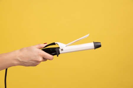 Closeup side view of woman hand holding showing iron curler, professional curling tongs, beauty care concept and hairstyling. Indoor studio shot isolated on yellow background.の写真素材