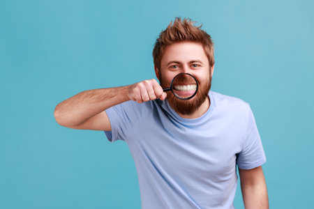 Portrait of funny positive young adult bearded man standing, holding magnifying glass on his teeth, looking at camera with toothy smile. Indoor studio shot isolated on blue background.の写真素材