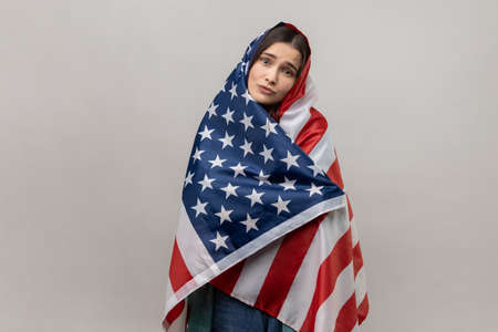 Portrait of relaxed young adult woman standing wrapped in USA flag, celebrating national holiday,...