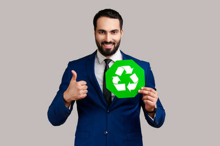Portrait of satisfied delighted bearded businessman holding pointing at green recycling sign, saving environment, wearing official style suit. Indoor studio shot isolated on gray background.の写真素材