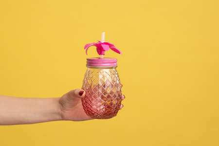 Closeup side view shot of woman hand holding pink glass jar mug with straw for cocktail or smoothie. Indoor studio shot isolated on yellow background.の写真素材