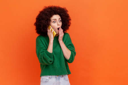 Astonished woman with Afro hairstyle wearing green casual style sweater talking via cell phone, hearing shocking news, covering mouth with palm. Indoor studio shot isolated on orange background.の写真素材