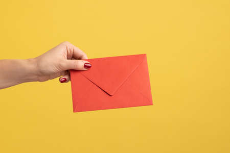 Profile side view closeup of woman hand holding red letter envelope, romantic message. Indoor studio shot isolated on yellow background.の写真素材