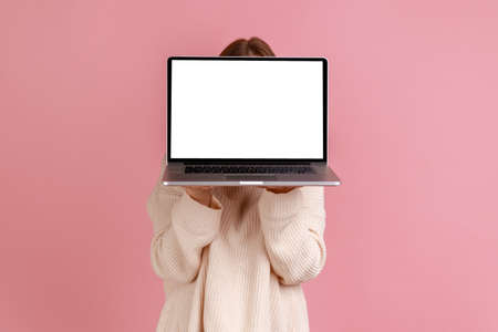 Portrait of blond woman holding laptop in hands, hiding her face behind notebook with empty display with copy space, wearing white sweater. Indoor studio shot isolated on pink background.の写真素材