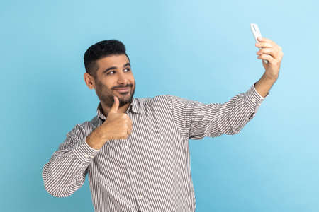 Satisfied man blogger showing thumbs up like gesture and winking looking at phone camera, making selfie or recording video, wearing striped shirt. Indoor studio shot isolated on blue background.の写真素材