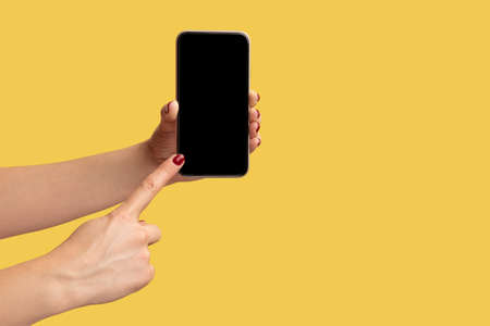 Profile side view closeup of woman hand holding and showing smart phone and pointing at empty display. Indoor studio shot isolated on yellow background.の写真素材