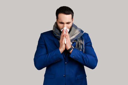Man wrapped in warm scarf sneezing in napkin, cleaning runny nose, suffering allergy or flu...