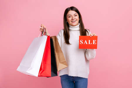 Satisfied female with dark hair holding sale card and shopping bags, huge discounts, low prices, wearing white casual style sweater. Indoor studio shot isolated on pink background.の写真素材