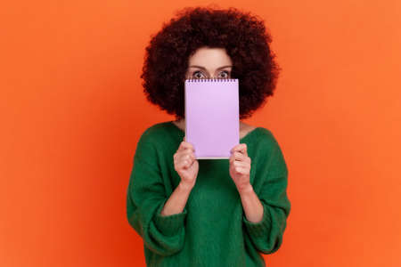 Portrait of woman with Afro hairstyle wearing green casual style sweater covering half of her face with paper organizer, looking at camera. Indoor studio shot isolated on orange background.の写真素材