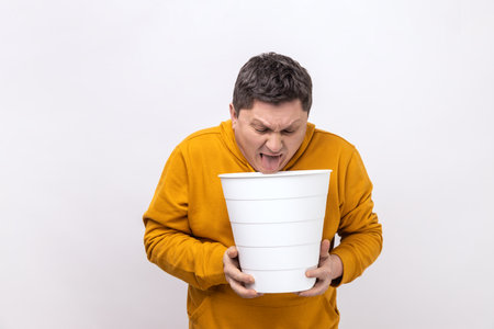 Food poisoning. Middle aged man feeling sudden stomach pain, suffering nausea and vomit, holding bin in hands, indigestion, wearing urban style hoodie. Indoor studio shot isolated on white background.の写真素材
