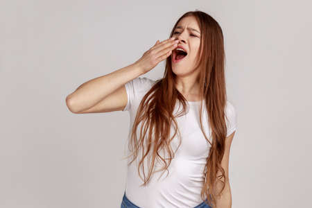 Portrait of attractive tired sleepy woman yawning, closing mouth with hand, exhausted with insomnia, lack of energy, wearing white T-shirt. Indoor studio shot isolated on gray background.の写真素材