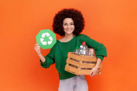 Smiling woman with Afro hairstyle wearing green casual style sweater holding box with plastic bottles and showing green recycling sign. Indoor studio shot isolated on orange background.の写真素材