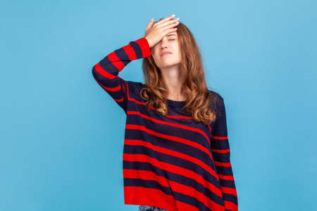 Portrait of unhappy girl standing with hand on head, feeling sorrow regret blaming herself for failure, wearing striped casual style sweater. Indoor studio shot isolated on blue background.の写真素材