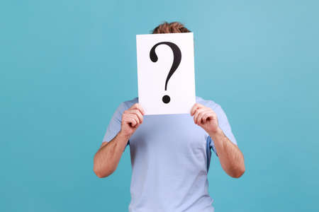 Portrait of unknown bearded man hiding his face behind white paper with question mark, finding smart solution, asking for advice. Indoor studio shot isolated on blue background.の写真素材