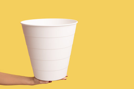 Profile side view closeup of woman hand holding white wastebasket, bin for garbage, reusable trash can. Indoor studio shot isolated on yellow background.の写真素材