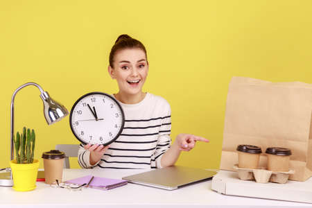 Happy optimistic woman office worker holding big wall clock pointing finger at paper bag, coffee...