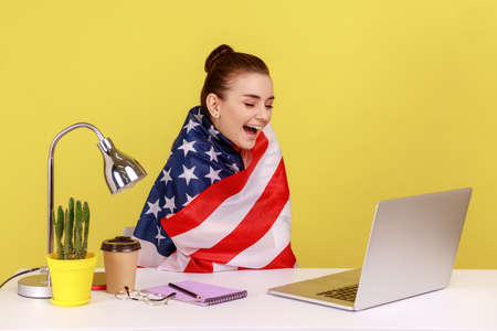 Happy woman employee sitting wrapped in USA flag, shouting for joy in office workplace, celebrating...