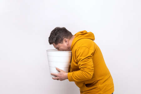 Side view of middle aged man feeling sudden stomach pain, suffering nausea and vomit, holding bin in hands, indigestion, wearing urban style hoodie. Indoor studio shot isolated on white background.の写真素材