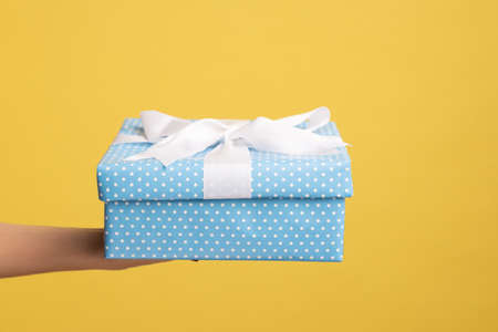Closeup side view of woman hand holding out blue gift box with ribbon, giving present on holiday, bonuses and surprises concept. Indoor studio shot isolated on yellow background.の写真素材