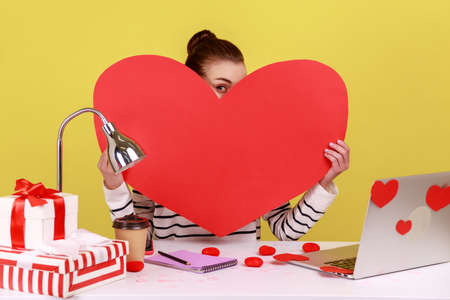 Portrait of shy woman sitting at workplace, holding and peeping from big red heart, expressing romantic feelings and love. Indoor studio studio shot isolated on yellow background.の写真素材