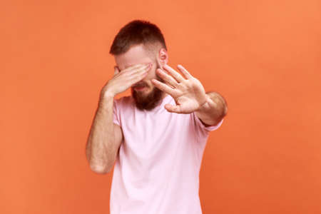 Portrait of man closing eyes with palm of hand, stretching out another hand forward touching space, looking for way in darkness, wearing pink T-shirt. Indoor studio shot isolated on orange background.の写真素材