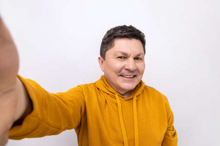Middle aged handsome man taking selfie, looking at camera POV and smiling, broadcasting livestream, point of view of photo, wearing urban style hoodie. Indoor studio shot isolated on white background.の写真素材