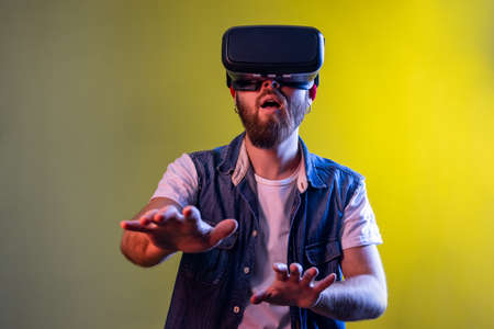 Amazed hipster man standing in virtual reality headset, playing video game and trying to touch something, wearing hat and denim vest. Indoor studio shot isolated on colorful neon light background.の写真素材
