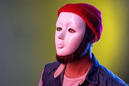 Side view of anonymous hipster man in white mask on face, hiding his identity, spying undercover, corruption, wearing hat and denim vest. Indoor studio shot isolated on colorful neon light background.の写真素材