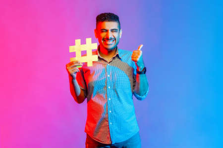 Portrait of man in shirt with toothy smile, holding hashtag symbol, showing thumb up, recommending...