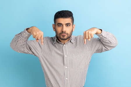 Here and now. Strict bearded handsome businessman pointing finger down, looking at camera with serious bossy facial expression, wearing striped shirt. Indoor studio shot isolated on blue background.の写真素材