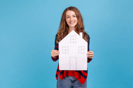 Smiling woman wearing striped casual style sweater, holding paper house, looking at camera with pleasant smile, dreaming of own home. Indoor studio shot isolated on blue background.の写真素材