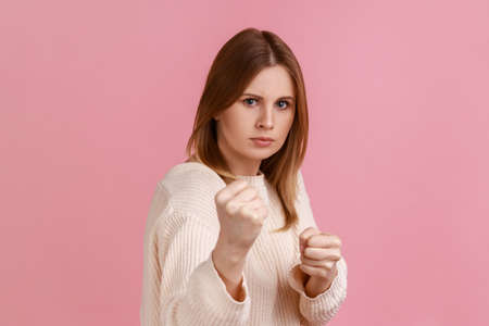 Portrait of angry confident attractive blond woman clenching fists, showing boxing gesture and ready to punch, wearing white sweater. Indoor studio shot isolated on pink background.の写真素材