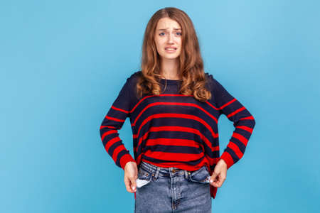 Im bankrupt. Unhappy poor woman, turning empty pockets inside out and looking frustrated by overspend, lack of money, wearing striped casual sweater. Indoor studio shot isolated on blue background.の写真素材