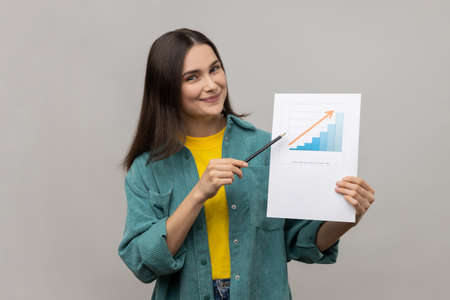 Portrait of positive businesswoman holding paper chart showing increase of workers salaries, pointing to paper, wearing casual style jacket. Indoor studio shot isolated on gray background.の写真素材