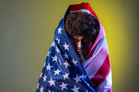 Portrait of hipster man with Afro hairstyle standing wrapped in american flag, looking away with funny facial expression and pout lips. Indoor studio shot isolated on colorful neon light background.の写真素材