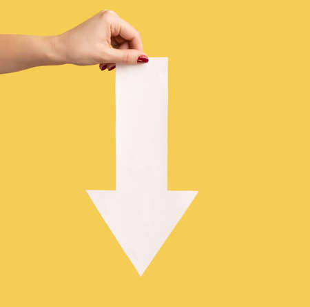 Closeup profile side view of woman hand showing white arrow pointing down, downgrade. Indoor studio shot isolated on yellow background.の写真素材