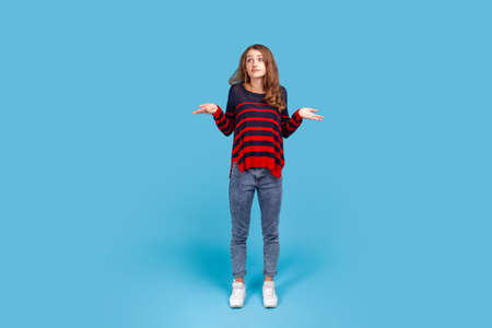 Full length portrait of uncertain woman wearing striped sweater looking desperate, making helpless gesture with her hands, doesnt know what to do. Indoor studio shot isolated on blue background.の写真素材