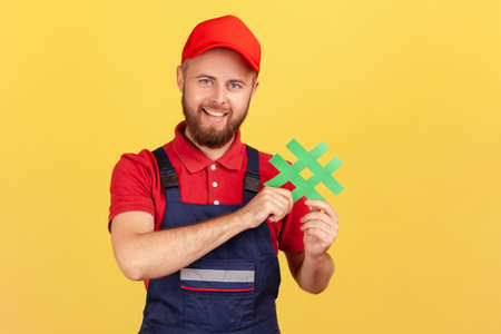 Portrait of positive delighted workman wearing blue overalls standing, holding green hashtag, looking at camera with happy expression. Indoor studio shot isolated on yellow background.の写真素材
