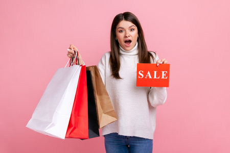 Astonished female with dark hair holding sale card and shopping bags, huge discounts, low prices,...