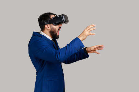 Side view of man in vr headset, playing virtual reality game with shocked facial expression, outstretching hands, wearing official style suit. Indoor studio shot isolated on gray background.の写真素材