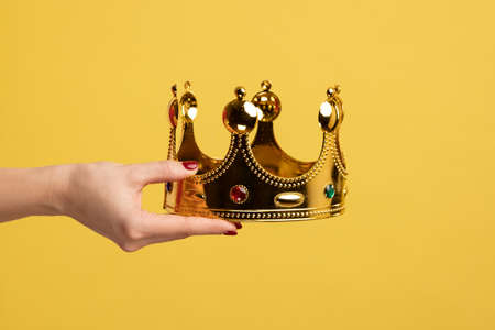 Closeup of woman hand holding golden crown, concept of awards ceremony, privileged status, superior position. Indoor studio shot isolated on yellow background.の写真素材