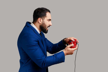 Profile portrait of concentrated attractive bearded man holding in hands red gamepad joystick,...