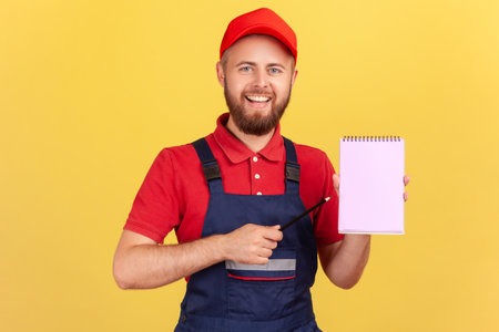 Optimistic smiling workman wearing blue overalls and red cap, holding pointing at paper notebook with pleasant smile, presenting copy space. Indoor studio shot isolated on yellow background.の写真素材